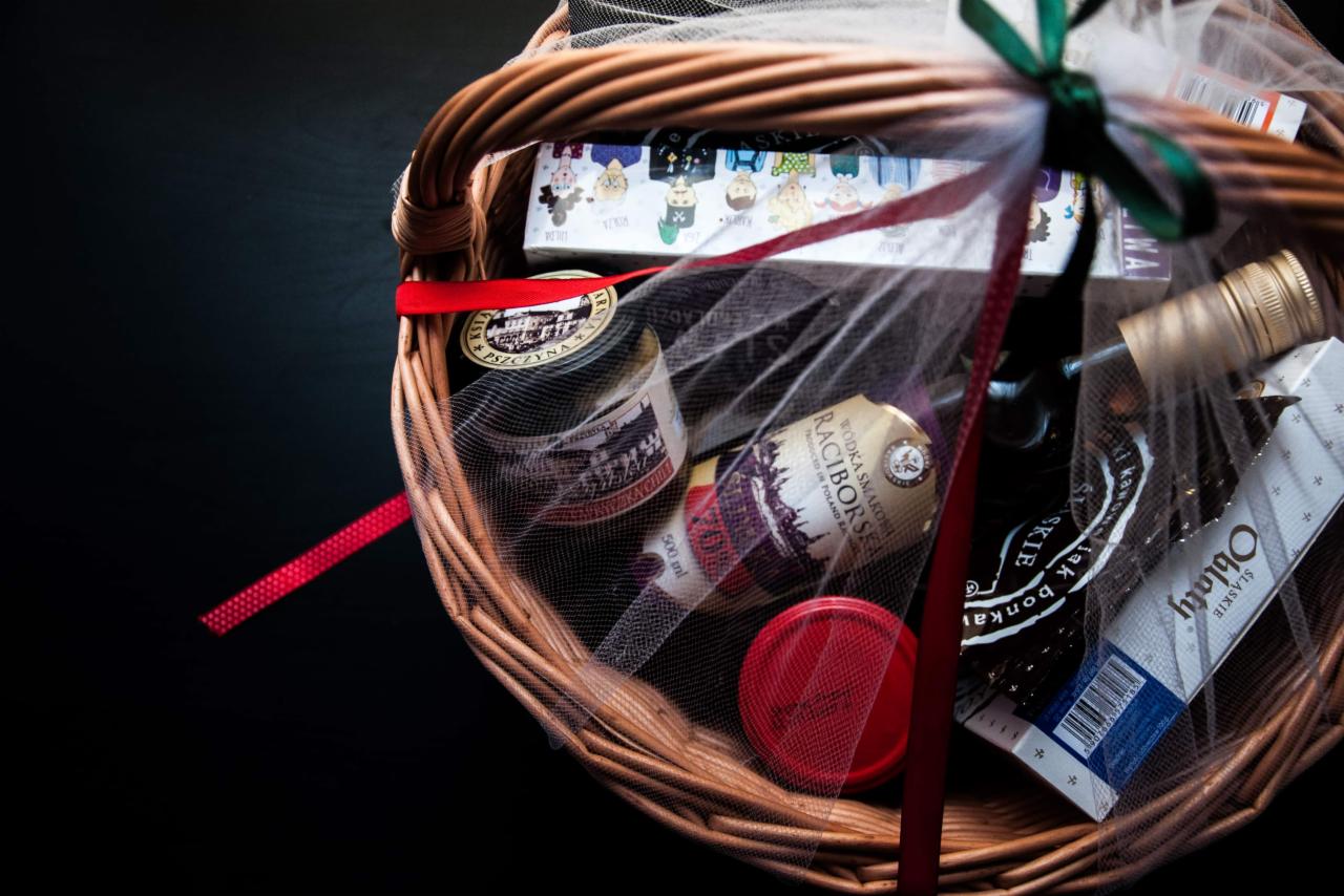 Wnętrze wiklinowego kosza, wypełnionego produktami spożywczymi i napojami alkoholowymi, opakowanego w białą siatkę i przewiązanego czerwoną wstążką oraz zieloną kokardą, widoczne etykiety z nazwami...
