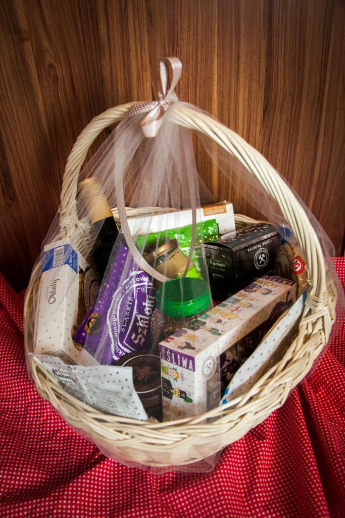 Wiklinowy kosz wypełniony różnorodnymi produktami spożywczymi, w tym słodyczami i napojami, zapakowany w tiul i ozdobiony kokardą, na tle drewnianej ściany i czerwonej tkaniny w białe kropki.
