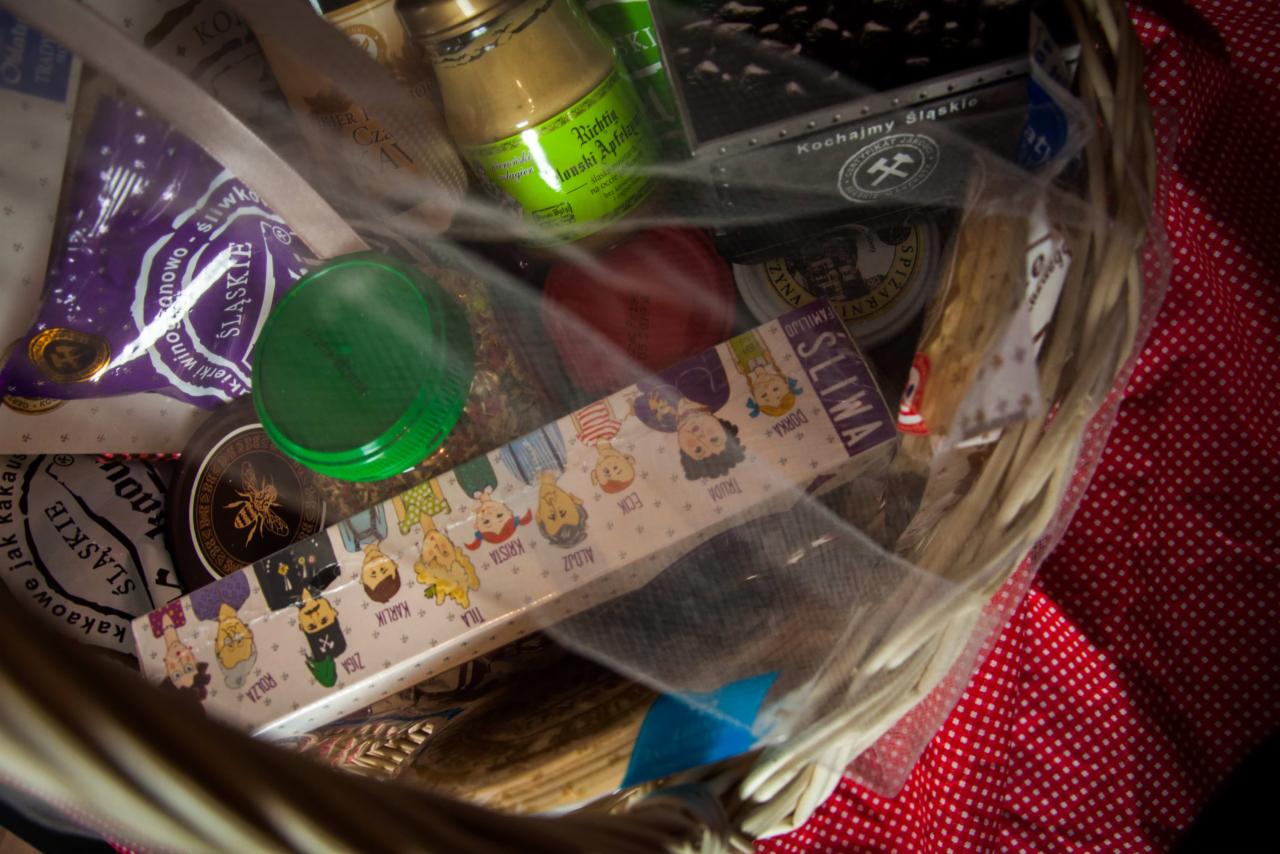 Zawartość wiklinowego kosza, widoczna przez przezroczyste opakowanie, zawiera regionalne śląskie produkty spożywcze, w tym słodycze, miód, musztardę i inne przysmaki, ułożone na czerwonej tkaninie...