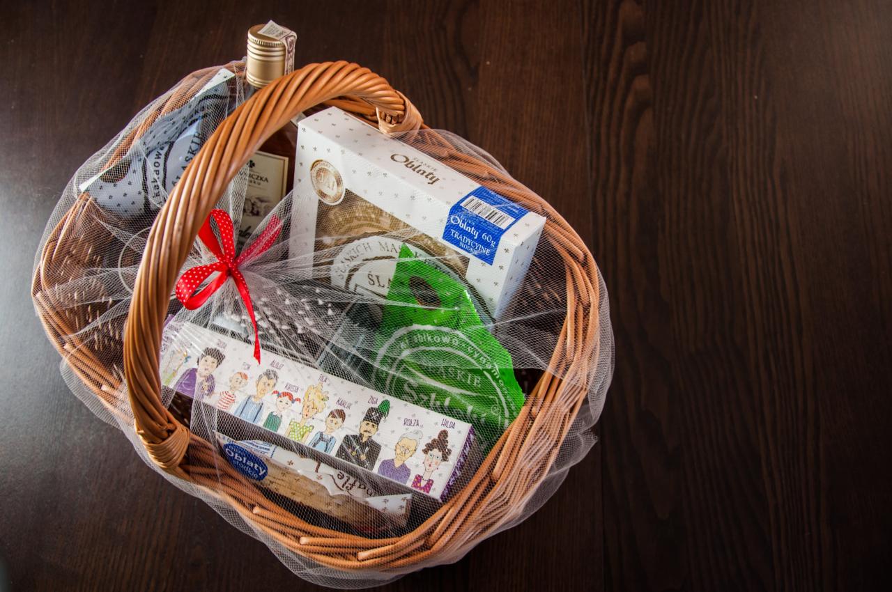 Wiklinowy kosz wypełniony produktami spożywczymi, w tym butelką alkoholu, wafle i słodycze, całość owinięta w tiul z czerwoną kokardką na ciemnym drewnianym tle.