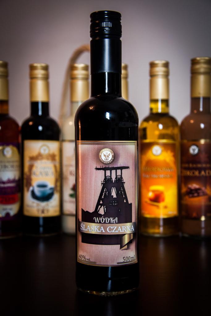 Czarna butelka wódki śląskiej z etykietą przedstawiającą szyb kopalni na tle innych butelek likierów i nalewek.