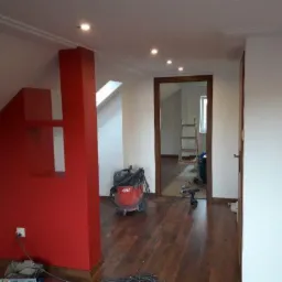 Wnętrze poddasza w trakcie prac: czerwona ściana z wnęką, drewniana podłoga, widoczny odkurzacz przemysłowy, drabina w przejściu do kolejnego pomieszczenia.