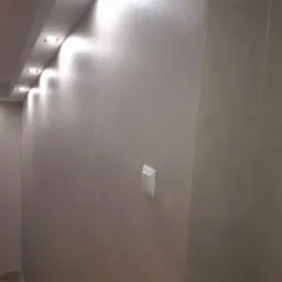 Ściana pokryta szarą gładzią w narożniku pomieszczenia, oświetlona punktowymi lampami sufitowymi, z widocznym włącznikiem światła.