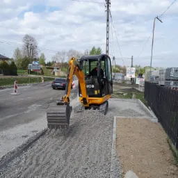 Żółta mini koparka JCB rozgarnia gruz na przygotowywanym podłożu chodnika z obrzeżami, w tle ulica z jadącym samochodem i słup elektryczny.