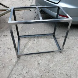 Surowa, spawana rama z metalowych profili o kwadratowym przekroju, stojąca na betonowej nawierzchni, z przyczepką i fragmentem samochodu w tle.