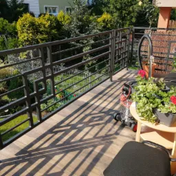 Nowoczesna balustrada metalowa na balkonie z drewnopodobną podłogą, widok na zadbany ogród, kwiaty w donicach na stoliku, odkurzacz do sprzątania balkonu.