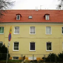 Dwupiętrowy budynek z żółtą elewacją i czerwonym dachem z trzema lukarnami, widoczny znak drogowy.