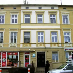 Odnowiona kamienica z żółtą i białą elewacją, widoczne okna z białymi ramami i detalami architektonicznymi, parter zajmują sklepy.