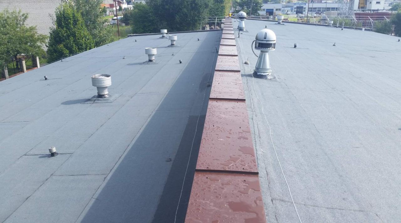 Płaski dach przemysłowy pokryty papą z licznymi kominkami wentylacyjnymi, z widocznymi śladami wilgoci i nowo zamontowaną obróbką blacharską w kolorze ceglastym.