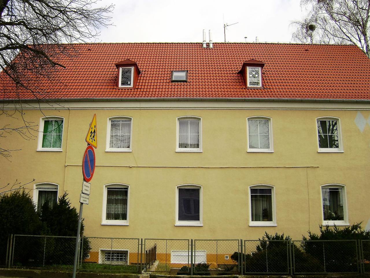 Dwupiętrowy budynek z żółtą elewacją i czerwonym dachem z trzema lukarnami, widoczny znak drogowy.