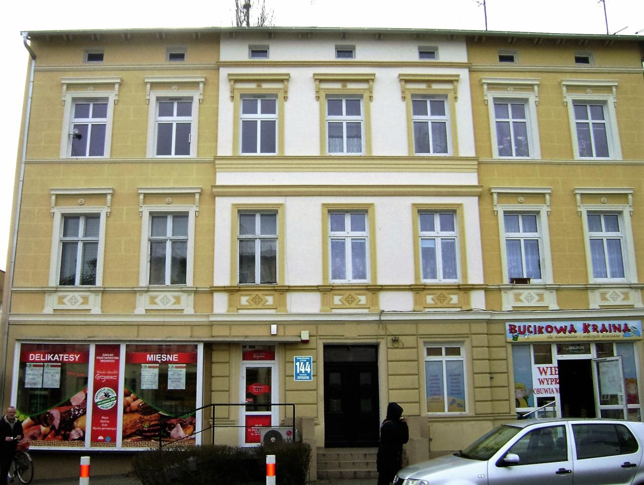 Odnowiona kamienica z żółtą i białą elewacją, widoczne okna z białymi ramami i detalami architektonicznymi, parter zajmują sklepy.