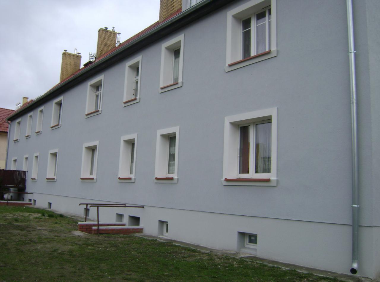 Długi budynek wielorodzinny z szarą elewacją, białymi obramowaniami okien i brązowymi parapetami, widoczne kominy i rynna spustowa.