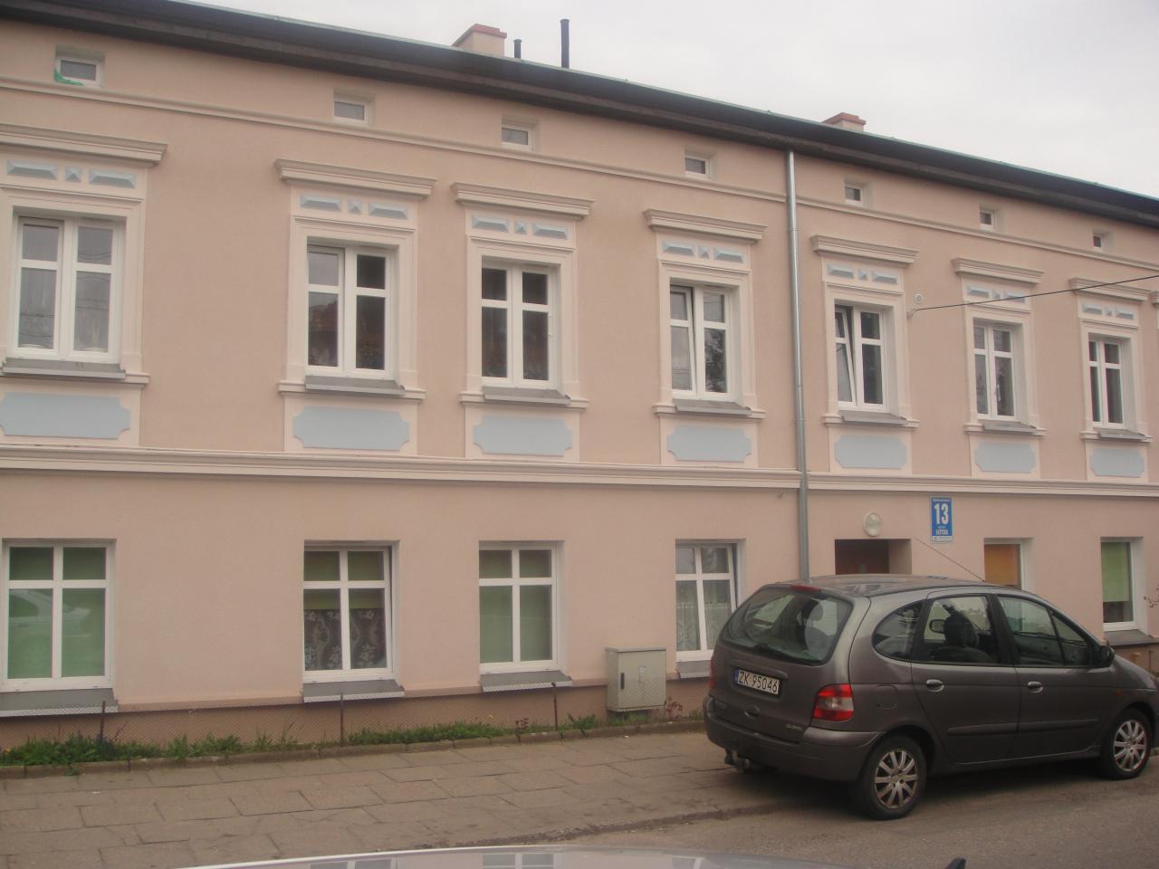 Odnowiona elewacja dwupiętrowego budynku mieszkalnego w pastelowym różu z białymi obramowaniami okien i dekoracyjnymi elementami, widoczny samochód osobowy zaparkowany przed budynkiem.