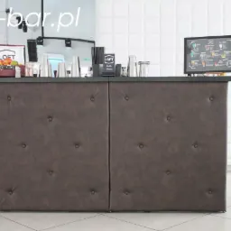 Frontowy widok baru z brązowym, pikowanym obiciem, na blacie szklanki, owoce w koszyku oraz menu drinków; w tle biała ściana z kwadratowych paneli i ciemne drzwi.