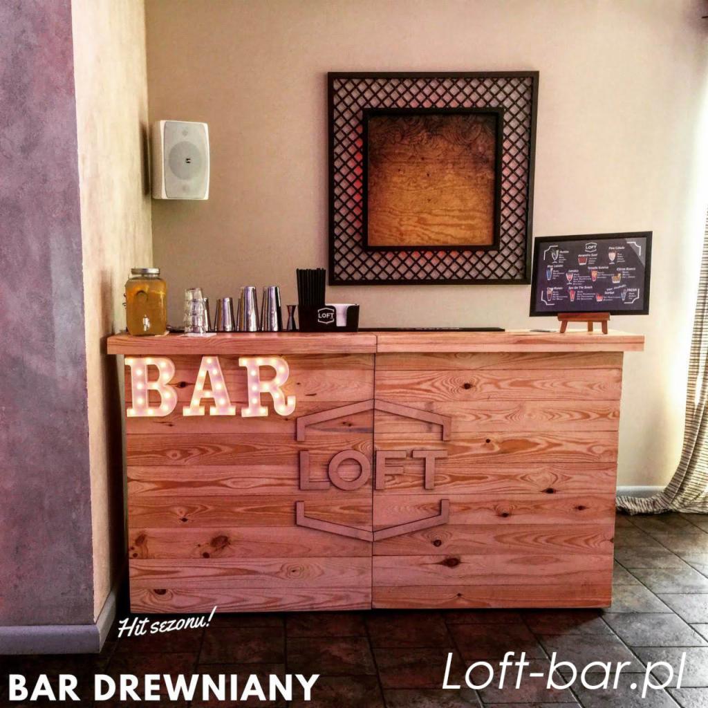 Drewniany bar z podświetlanym napisem 'BAR' i logo 'LOFT', uzupełniony o szklanki, shaker, menu drinków i dekoracje ścienne.