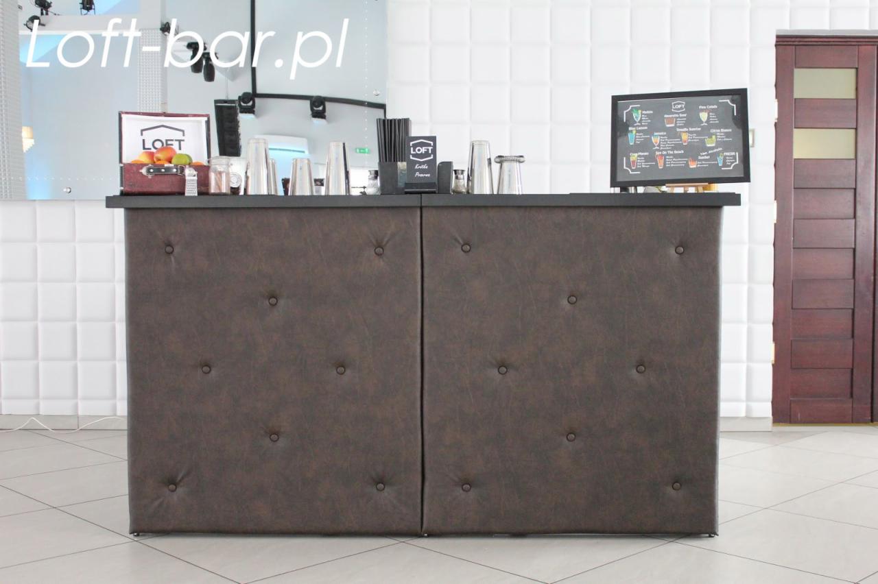 Frontowy widok baru z brązowym, pikowanym obiciem, na blacie szklanki, owoce w koszyku oraz menu drinków; w tle biała ściana z kwadratowych paneli i ciemne drzwi.