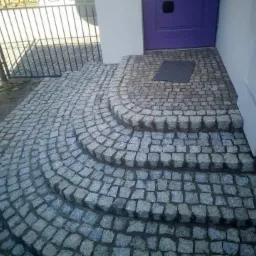 Granitowe schody wejściowe ułożone w półokrągły wzór, prowadzące do fioletowych drzwi wejściowych z wycieraczką, widok z góry.