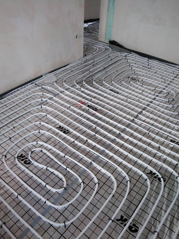 Układ ogrzewania podłogowego z białych rur na czarnej siatce, widoczne ściany w surowym stanie, w tle kontynuacja instalacji w kolejnym pomieszczeniu.