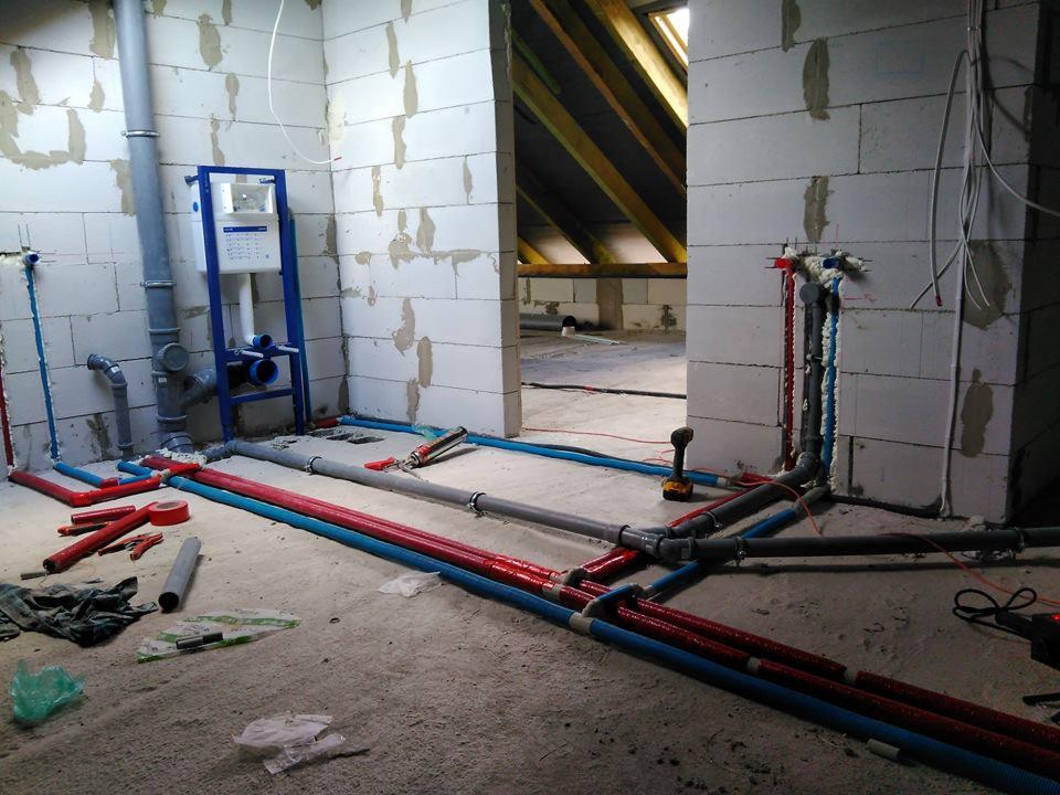 Wnętrze w trakcie montażu instalacji hydraulicznej w surowym stanie, widoczne rury w kolorach niebieskim i czerwonym prowadzone po podłodze, stelaż podtynkowy WC z niebieską ramą, ściany z betonu...