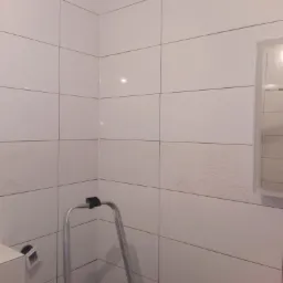 Ściana łazienki wyłożona białymi płytkami z poziomym pasem dekoracyjnym, widoczna drabina aluminiowa i częściowo lustro, w trakcie prac wykończeniowych.