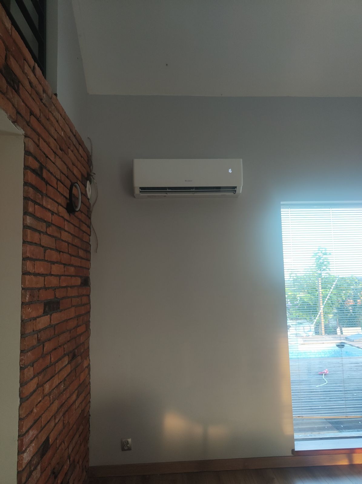 Biała klimatyzacja ścienna zamontowana w pomieszczeniu z ceglaną ścianą i widokiem na basen przez okno z żaluzjami.