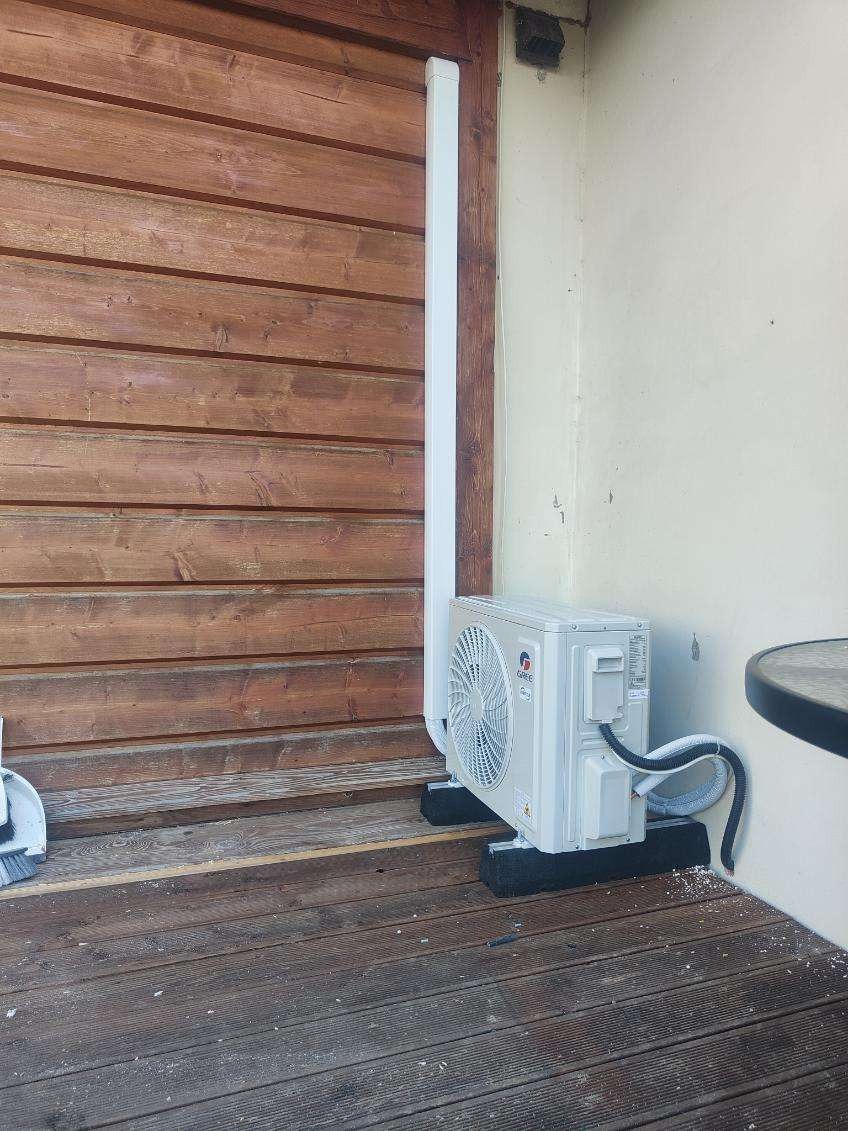 Zewnętrzna jednostka klimatyzacji marki Gree zamontowana na czarnych podkładkach na drewnianym tarasie, z widoczną białą rurą odprowadzającą i fragmentem okrągłego stolika.