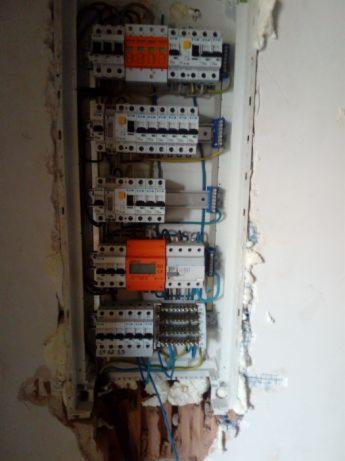 Rozdzielnica elektryczna z zamontowanymi bezpiecznikami i licznikiem energii, widoczne nieestetyczne wykończenie ściany wokół skrzynki.