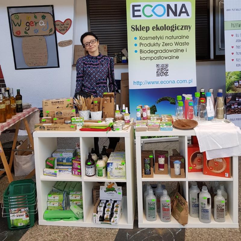 Stoisko z ekologicznymi środkami czystości i kosmetykami naturalnymi, prezentowane na białych regałach; kobieta w okularach stoi za stoiskiem, obok baneru reklamowego firmy ECONA.