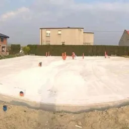 Wylana płyta fundamentowa na działce z widocznymi wystającymi rurami instalacyjnymi, w tle domy w różnym stanie budowy i zabudowania gospodarcze.