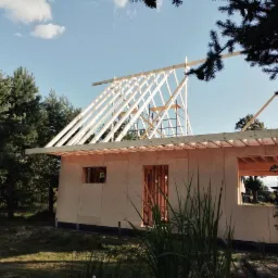 Wznoszenie konstrukcji szkieletowej domu jednorodzinnego z widocznym drewnianym szkieletem dachu, oszalowaną płytami OSB ścianą oraz rusztowaniem w tle, na tle zieleni drzew i błękitnego nieba.
