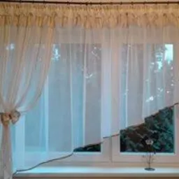 Delikatna, kremowa firanka woalowa z marszczeniem na karniszu, asymetrycznie podpięta, odsłaniająca widok na zieleń za oknem. Na parapecie mała, dekoracyjna roślina.