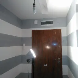 Korytarz z pomalowanymi ścianami w poziome pasy szaro-białe, oświetlony lampą budowlaną, widoczna drewniana brama wejściowa i wisząca żarówka.