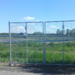 Dwu-skrzydłowa brama z siatki stalowej, element ogrodzenia autostradowego, widoczna z perspektywy gruntu, z autostradą i lasem w tle pod błękitnym niebem.