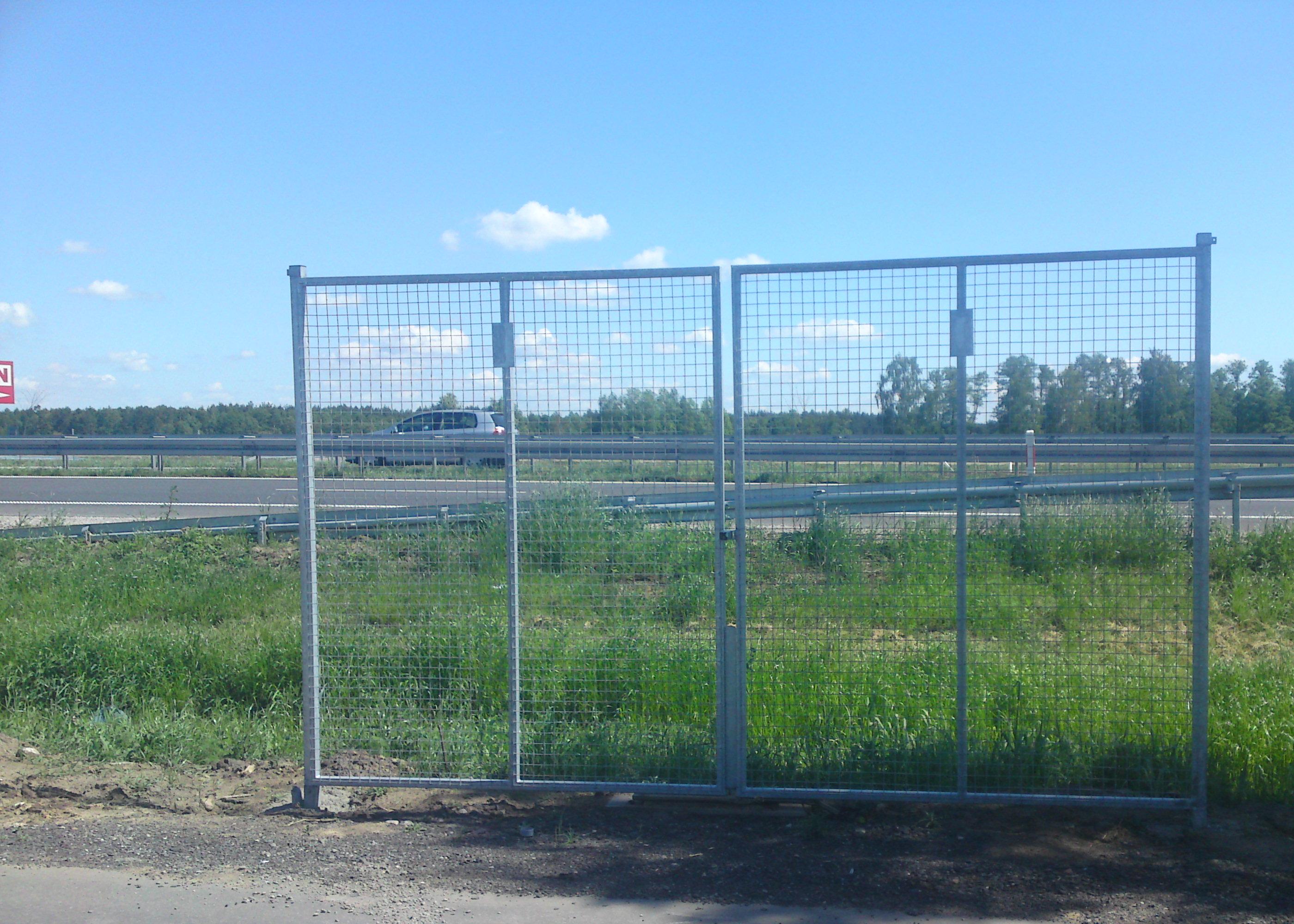 Dwu-skrzydłowa brama z siatki stalowej, element ogrodzenia autostradowego, widoczna z perspektywy gruntu, z autostradą i lasem w tle pod błękitnym niebem.