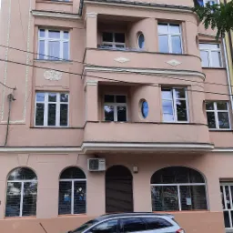 Trzypiętrowa kamienica z balkonami o pastelowej elewacji z okrągłymi oknami, widoczny fragment zaparkowanego samochodu.