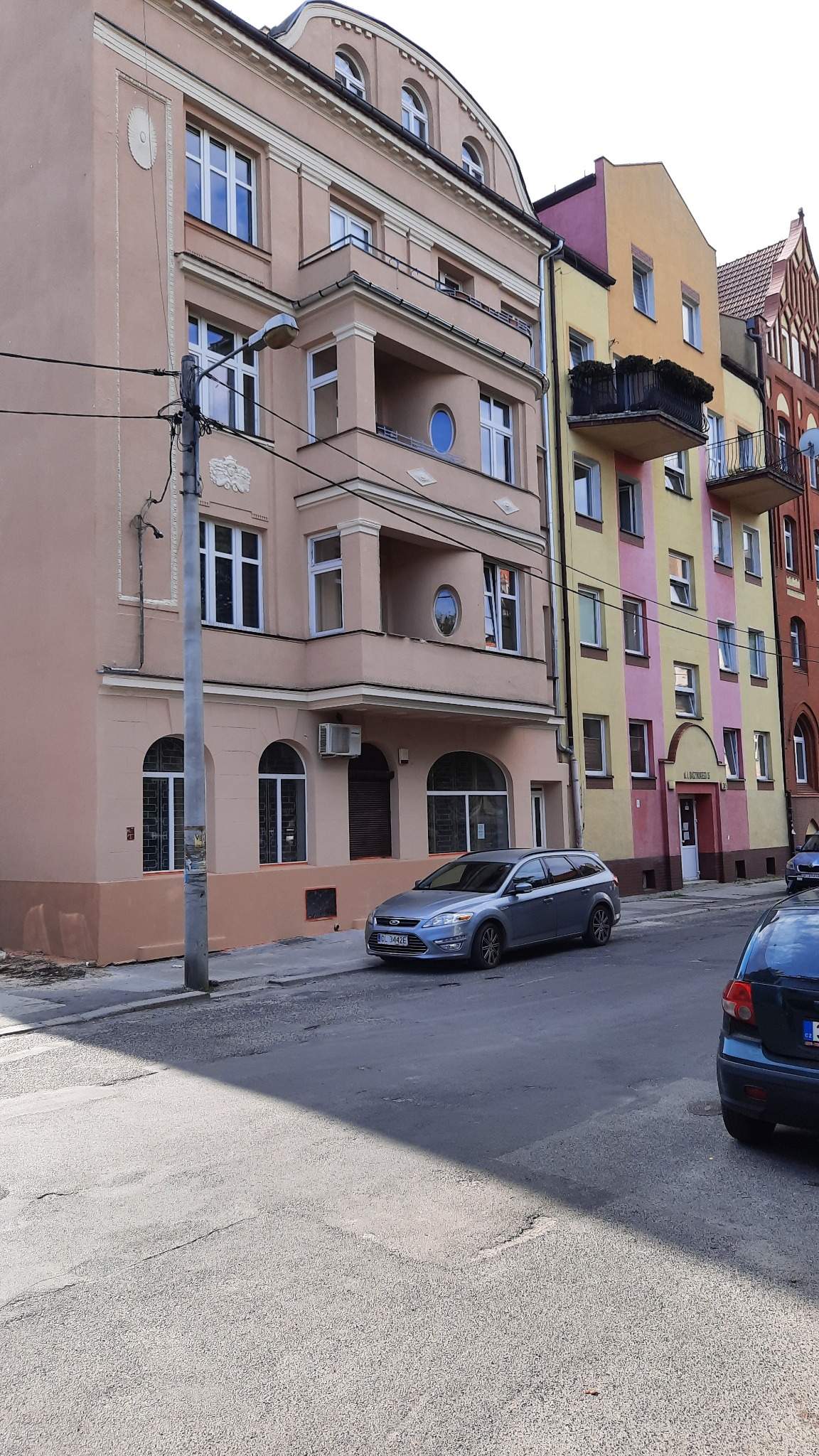 Kamienica z beżową elewacją, balkonami z okrągłymi oknami i ozdobnymi detalami, obok kolorowej kamienicy w odcieniach żółci i różu, na ulicy zaparkowany szary samochód.