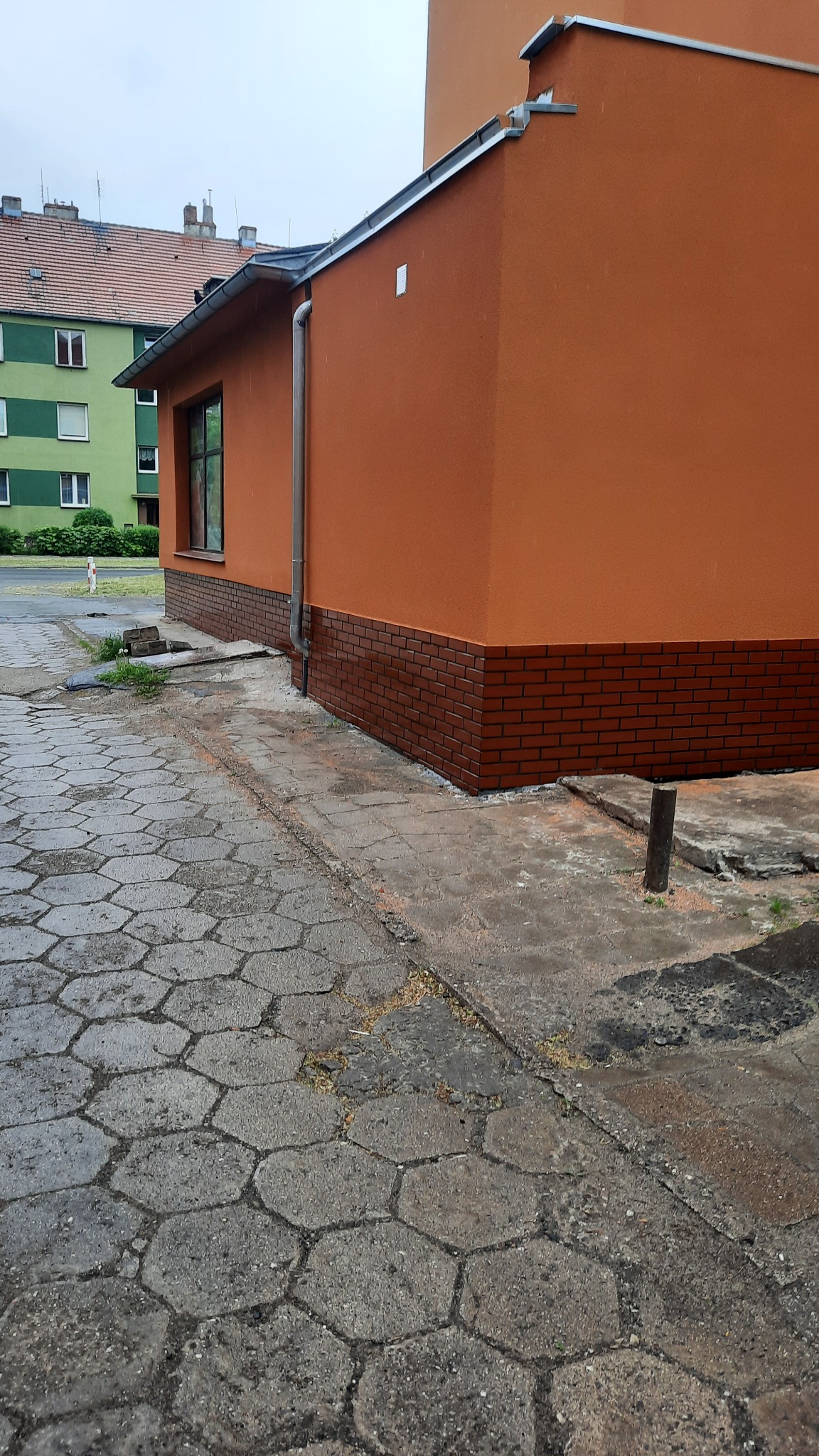 Narożnik budynku z nową, pomarańczową elewacją i czerwoną ceglaną podmurówką, widok z poziomu chodnika z szarej kostki brukowej.