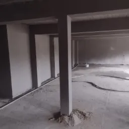 Wnętrze surowego budynku po nałożeniu szarego tynku maszynowego na ściany i filary, widoczne przewody na podłodze i wiadro z zaprawą, sufit betonowy.