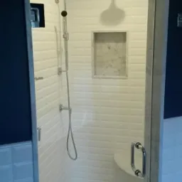 Nowoczesna kabina prysznicowa z białymi, wypukłymi płytkami, szklanymi drzwiami i marmurową wnęką na kosmetyki.