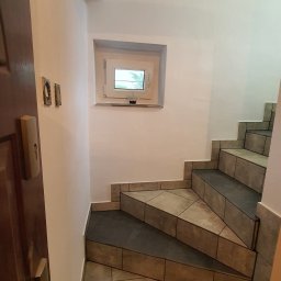 PLESS-FLIZ glazurnictwo,wykonczenia wnetrz Adrian Hlubek - Klatka schodowa z wyłożonymi płytkami schodami i małym oknem z widokiem na zieleń, widoczne ślady po starych kontaktach na ścianie, częściowo otwarte drewniane drzwi z mosiężną klamką.