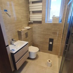 PLESS-FLIZ glazurnictwo,wykonczenia wnetrz Adrian Hlubek - Jasna łazienka z beżowymi płytkami imitującymi kamień, wisząca szafka z umywalką, podwieszana toaleta, grzejnik drabinkowy, kabina prysznicowa ze szklanymi drzwiami, oraz butelki kleju i pianki...