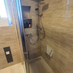 PLESS-FLIZ glazurnictwo,wykonczenia wnetrz Adrian Hlubek - Beżowa kabina prysznicowa z deszczownicą, półką na kosmetyki w ścianie, składanym siedziskiem i rolką papieru toaletowego na podłodze.