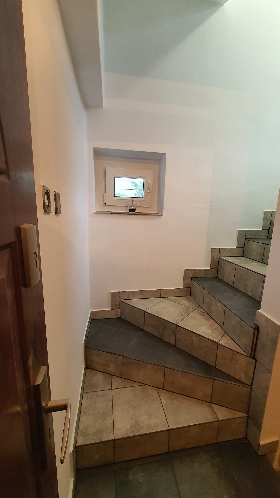 Klatka schodowa z wyłożonymi płytkami schodami i małym oknem z widokiem na zieleń, widoczne ślady po starych kontaktach na ścianie, częściowo otwarte drewniane drzwi z mosiężną klamką.