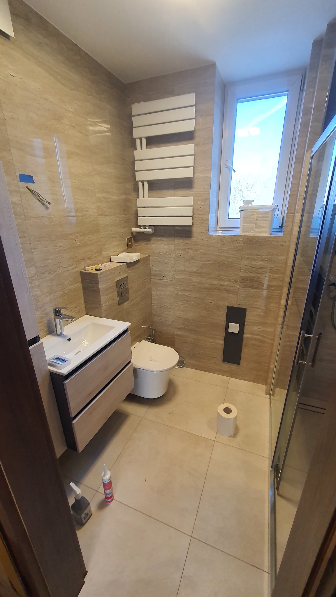 Jasna łazienka z beżowymi płytkami imitującymi kamień, wisząca szafka z umywalką, podwieszana toaleta, grzejnik drabinkowy, kabina prysznicowa ze szklanymi drzwiami, oraz butelki kleju i pianki...