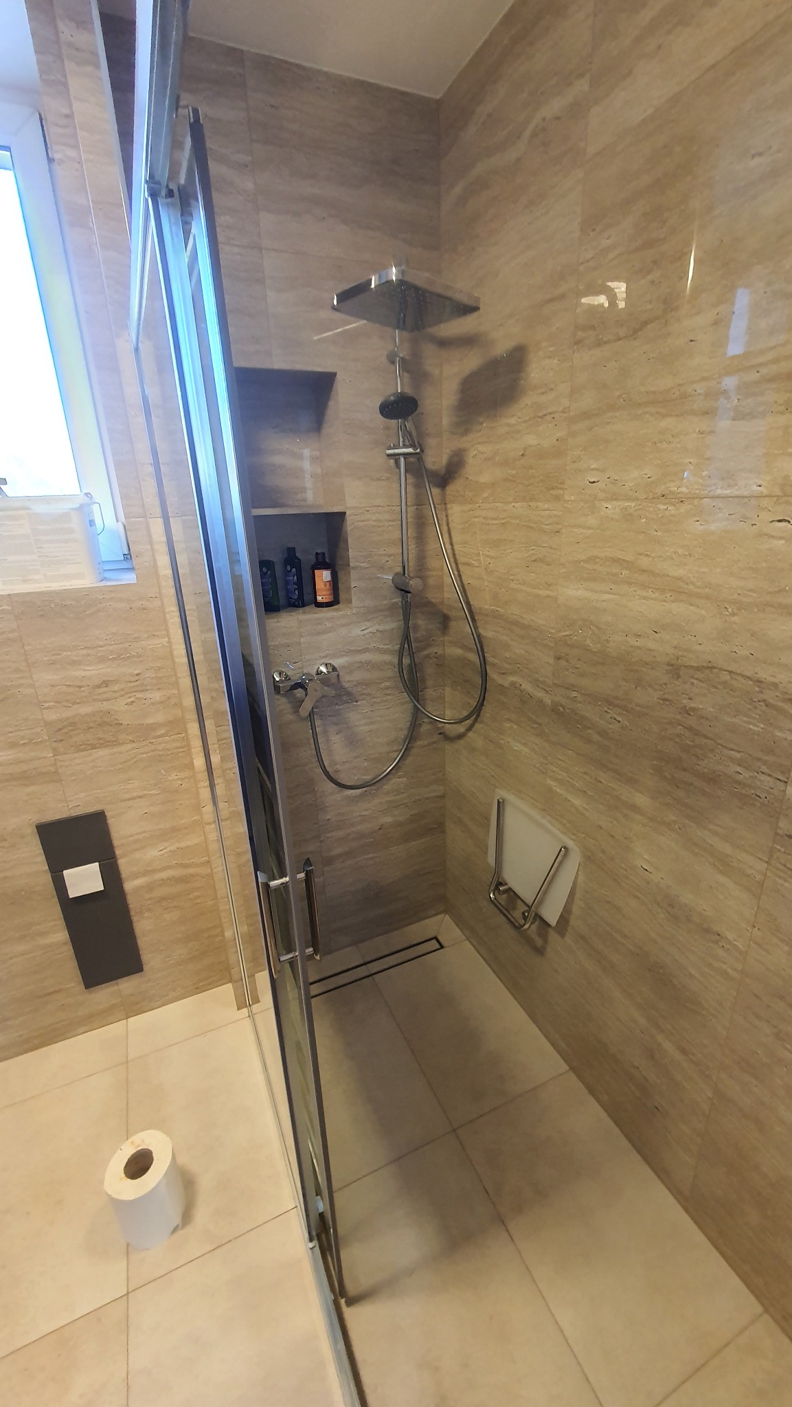 Beżowa kabina prysznicowa z deszczownicą, półką na kosmetyki w ścianie, składanym siedziskiem i rolką papieru toaletowego na podłodze.