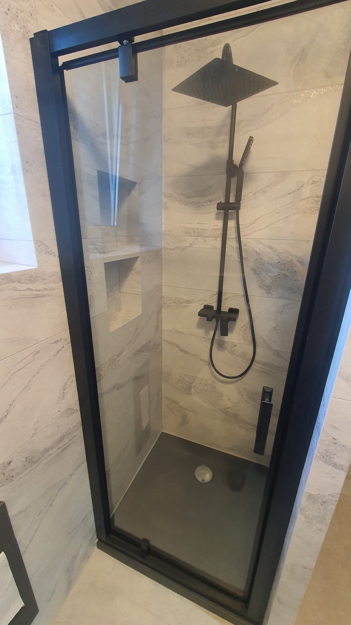 Nowoczesna kabina prysznicowa z czarną armaturą i ramą, marmurowe płytki na ścianach, odpływ liniowy w ciemnym brodziku, wnęka w ścianie.