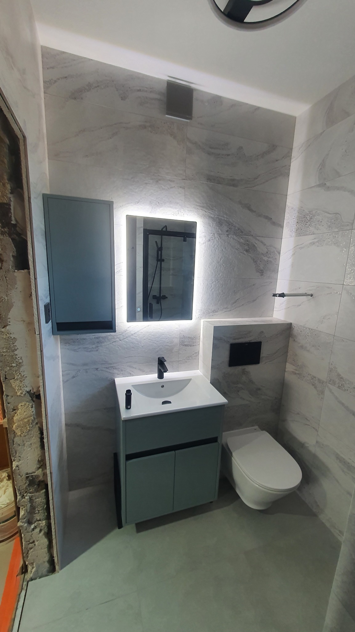 Nowoczesna łazienka z szarymi płytkami imitującymi marmur, umywalką z szafką w kolorze gołębim, podświetlanym lustrem i toaletą wiszącą; widoczny fragment ściany przed wykończeniem.