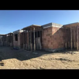 Surowe, ceglane ściany w trakcie budowy szeregu domów jednorodzinnych, z drewnianymi stemplami podtrzymującymi nadproża okienne, na piaszczystym terenie i pod błękitnym niebem.