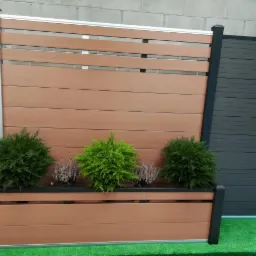 Panel ogrodzeniowy z kompozytu drewna w kolorze brązowym i antracytowym, z donicą i nasadzeniami iglaków i wrzosów, na tle ściany z szarego betonu.