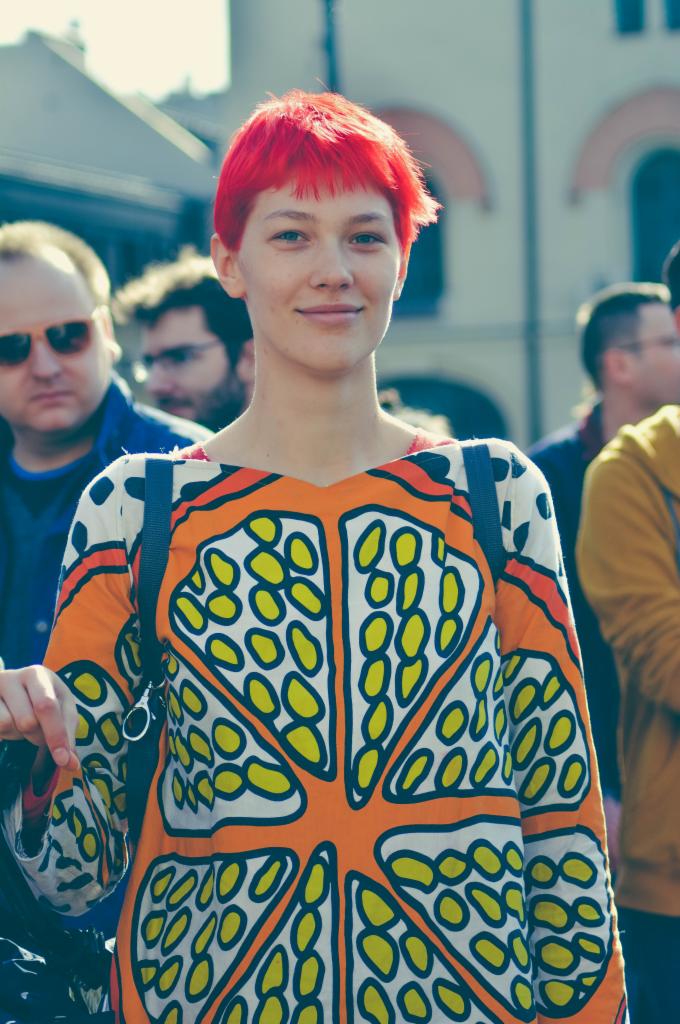 Portret kobiety z krótkimi, intensywnie czerwonymi włosami, ubranej w wzorzystą sukienkę w odcieniach pomarańczy, bieli i żółci, na tle miejskim z innymi osobami.